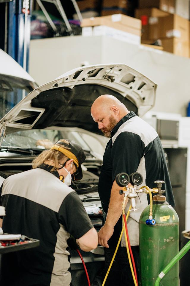 Two technicians servicing a vehicle's air conditioning with a gauge set and gas cylinder