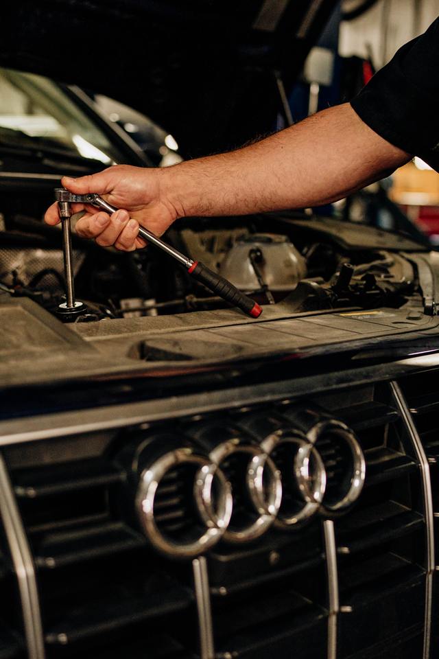 Technician using a ratchet in the engine bay of a black Audi, above the four-ring grille badge