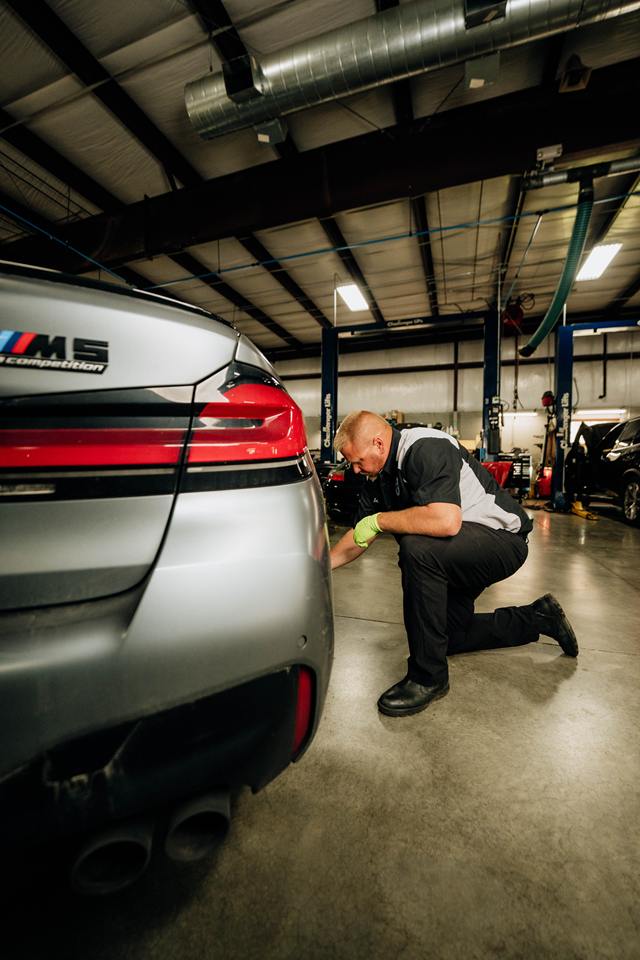 Technician kneeling to inspect the rear wheel of a silver BMW M5 Competition