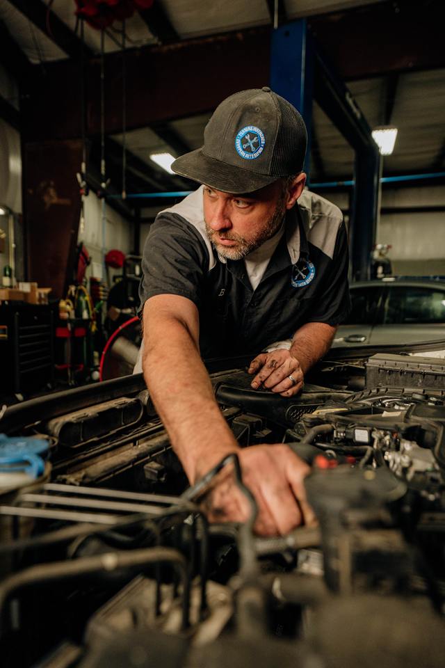 Technician in a Turning Wrenches cap reaching into an engine bay