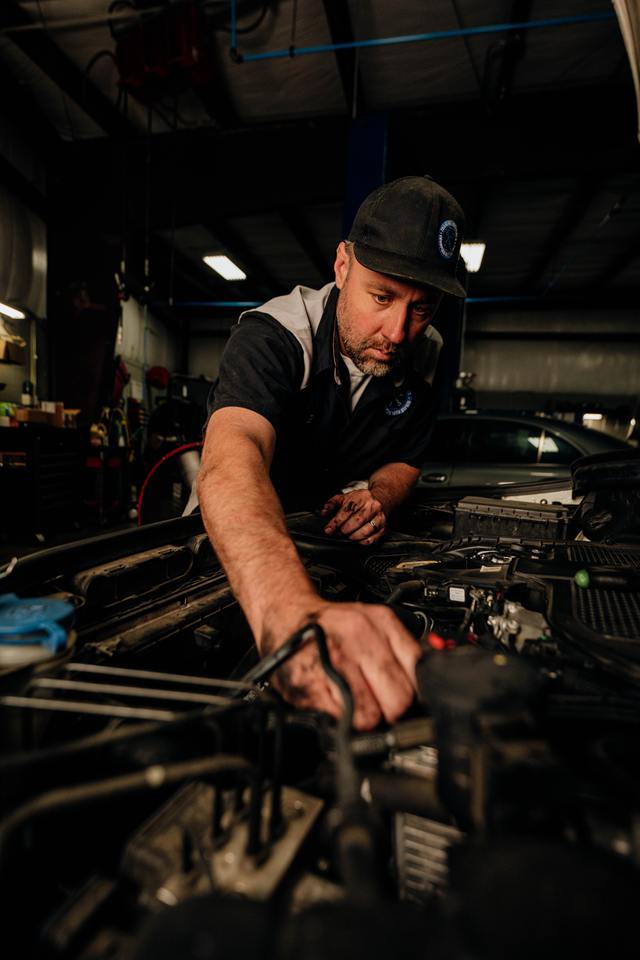 Technician reaching into an engine bay, with a gray sedan in the background