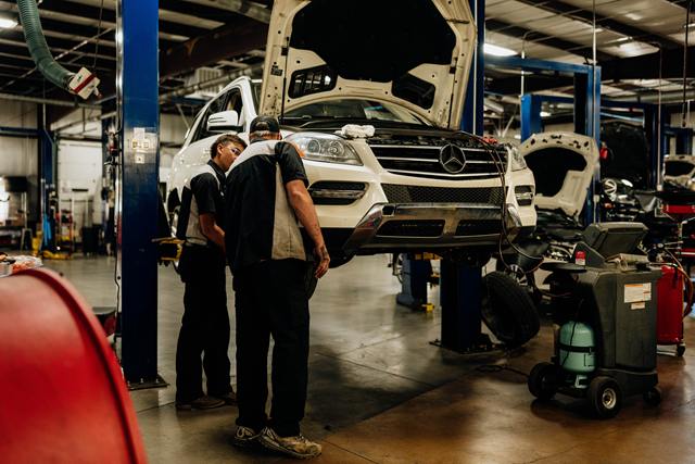 Two technicians inspecting a white Mercedes-Benz SUV raised on a lift