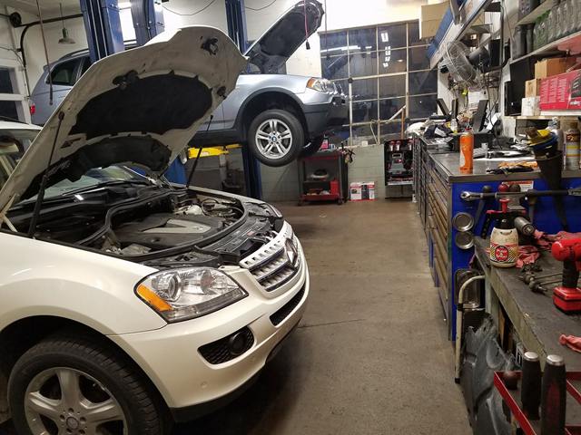 White Mercedes-Benz SUV with the hood open beside a workbench, with another SUV on the lift behind it