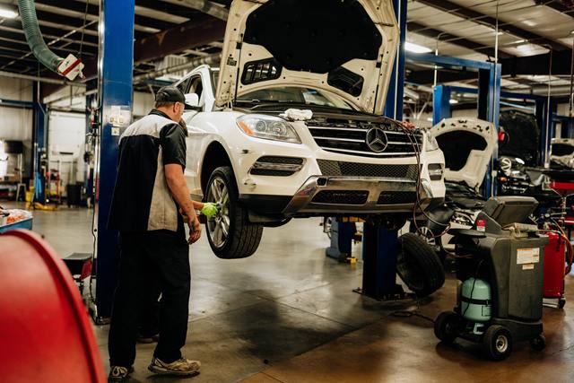 Technician servicing a white Mercedes-Benz SUV raised on a lift with the hood open