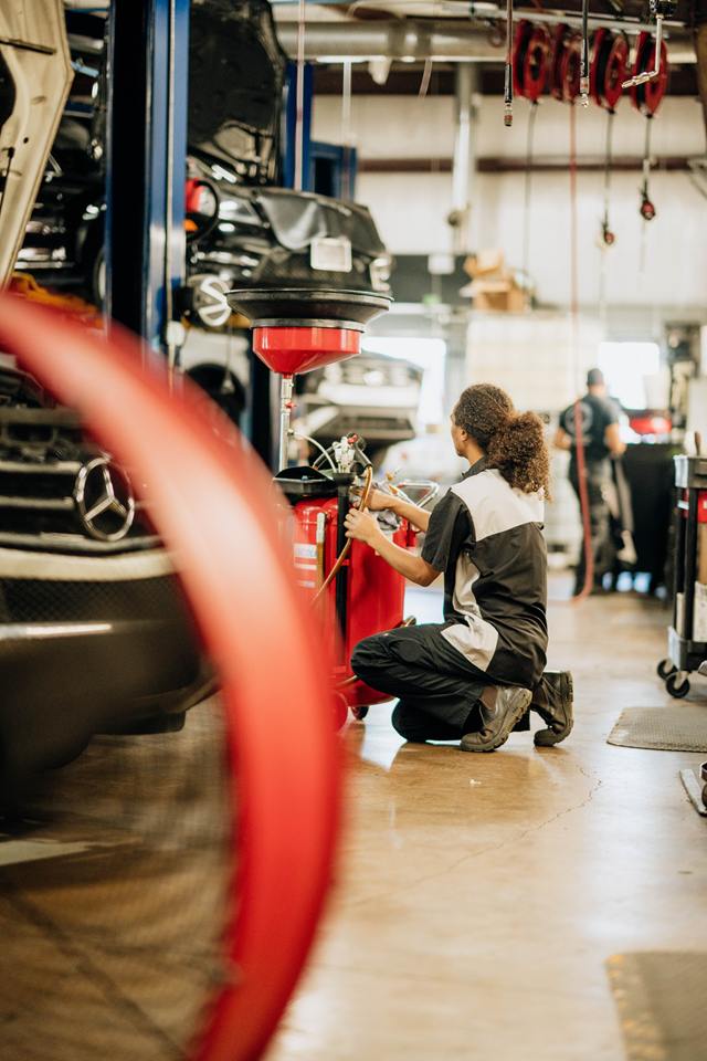 Technician kneeling at an oil drain beside a Mercedes-Benz on the lift